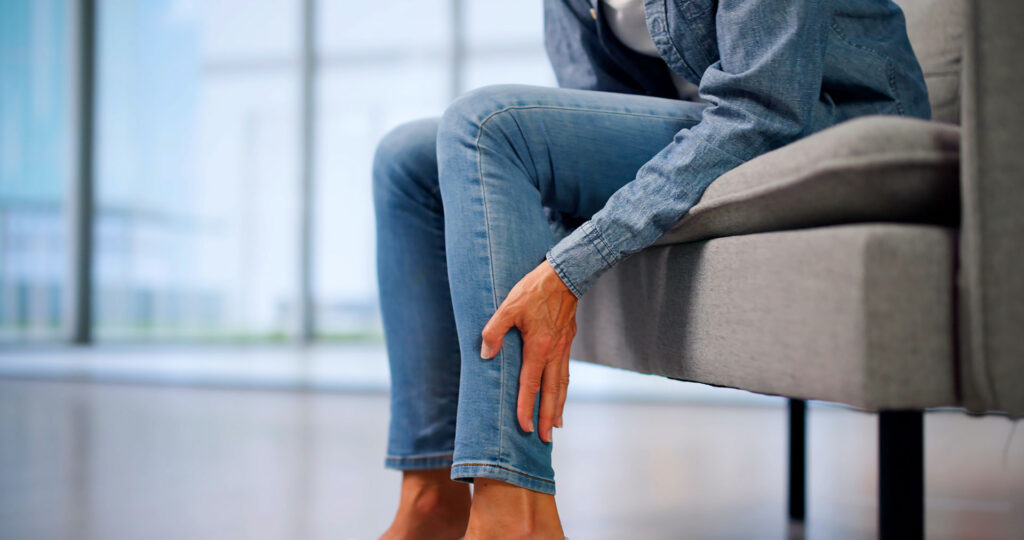 A person sitting on a gray couch wearing blue jeans and gently holding their lower leg where spider veins may develop.