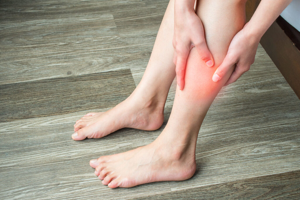 A close-up of a person sitting on a floor and holding their lower leg, with a reddish highlight indicating discomfort often associated with conditions like spider veins.