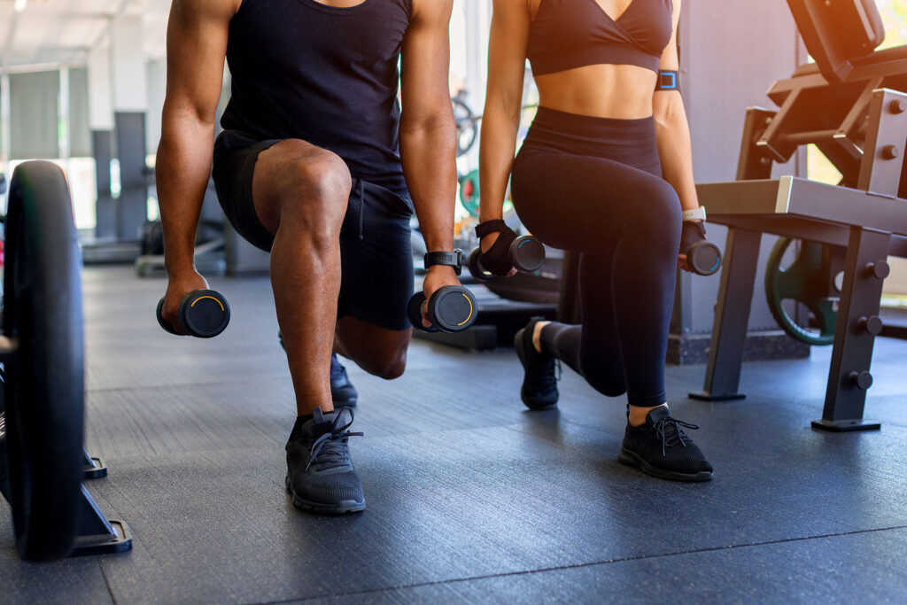 Man and woman performing weighted lunges in a gym setting to discuss high-impact training and varicose veins from working out.