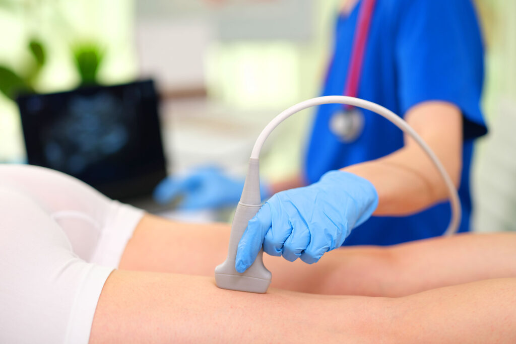Healthcare provider in blue scrubs performing a vascular ultrasound examination on a patient's leg using a transducer probe.