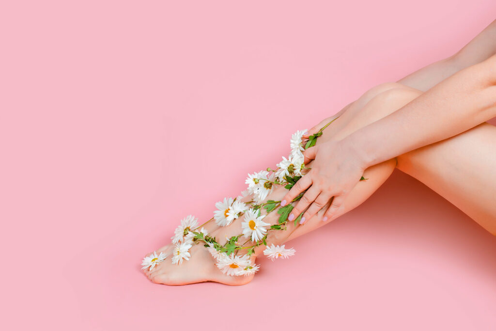 Woman sitting against a pink background resting white daisy flowers along her smooth legs, representing natural care and cream varicose vein remedies.
